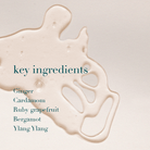 A close-up of a glossy beige liquid splash on a light warm background, with the text 'key ingredients' and a list reading Ginger, Cardamom, Ruby grapefruit, Bergamot, and Ylang Ylang in dark teal.