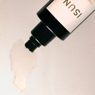 A close-up of a cosmetic bottle with a black pump dispensing a translucent, pale oil onto a light beige surface. The bottle label is white with black text, partially showing the brand name 'ISUN'.